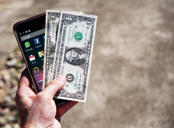 A picture of a hand holding a smart phone and a couple of dollar bills.